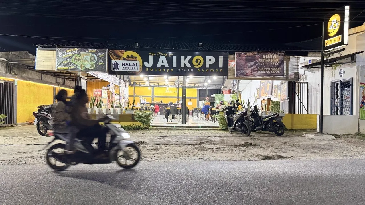 Tampak depan Jaikopi pada malam hari, spanduk hitam JAIKOPI dengan lampu menyala dan dinding kuning