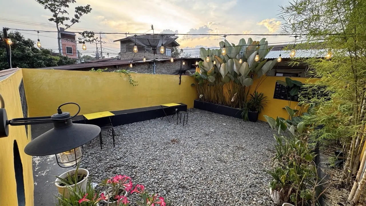 Area outdoor Jaikopi dengan dinding kuning, kerikil, tanaman, dan lampu gantung saat sore