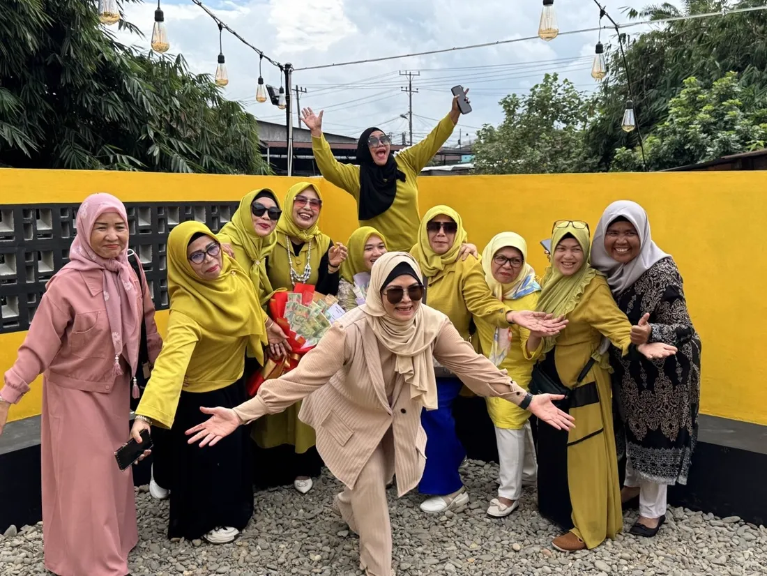 Rombongan ibu-ibu berbaju kuning merayakan dengan buket di area outdoor dinding kuning